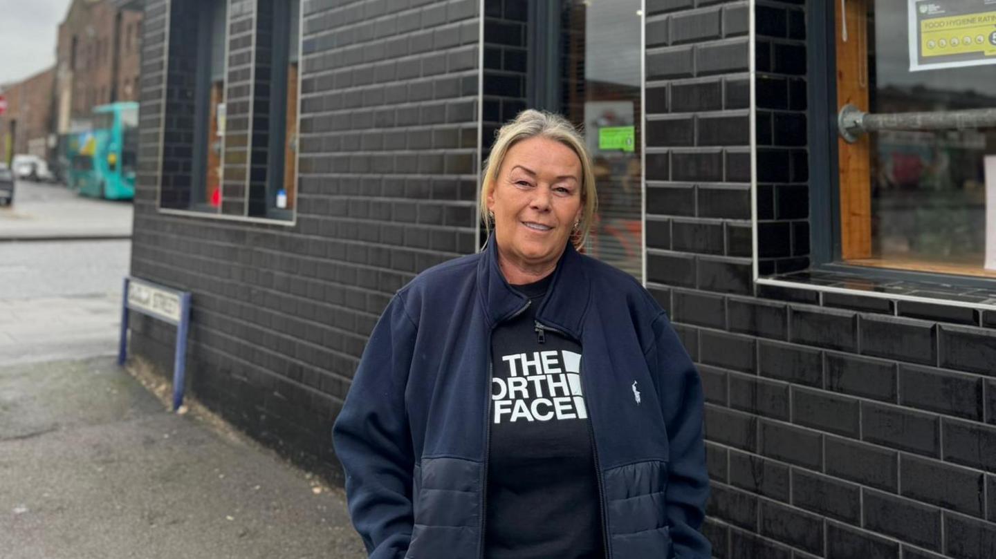 Marie Williams, who has blonde hair and is wearing a blue fleece over a black North Face branded t-shirt, stands in front of Stanleys Cafe which has black tiles installed on the brick-work