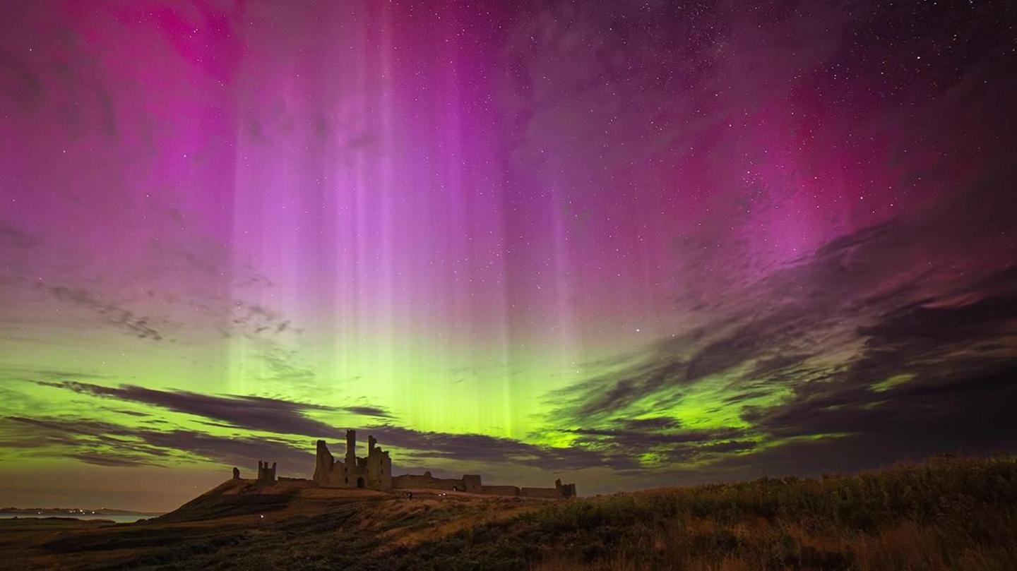 The night sky filled with bright purple and shafts of white light fills the top of the image while greens of the aurora are seen closer to the horizon where a rolling hill with a ruined castle sits.