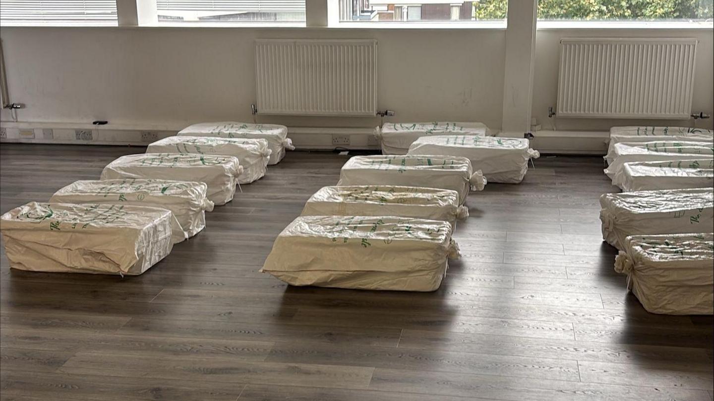 Three rows of boxes covered in white plastic bags, laid out on a bare wooden floor in a large room