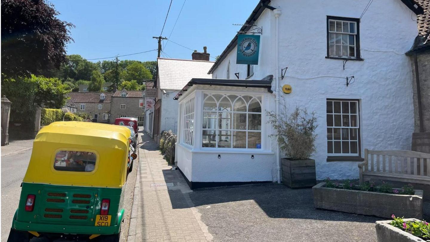 A picturesque village street, with a whitewashed cottage-type building, a business with the hanging sign on the side saying Bluebird Cafe.