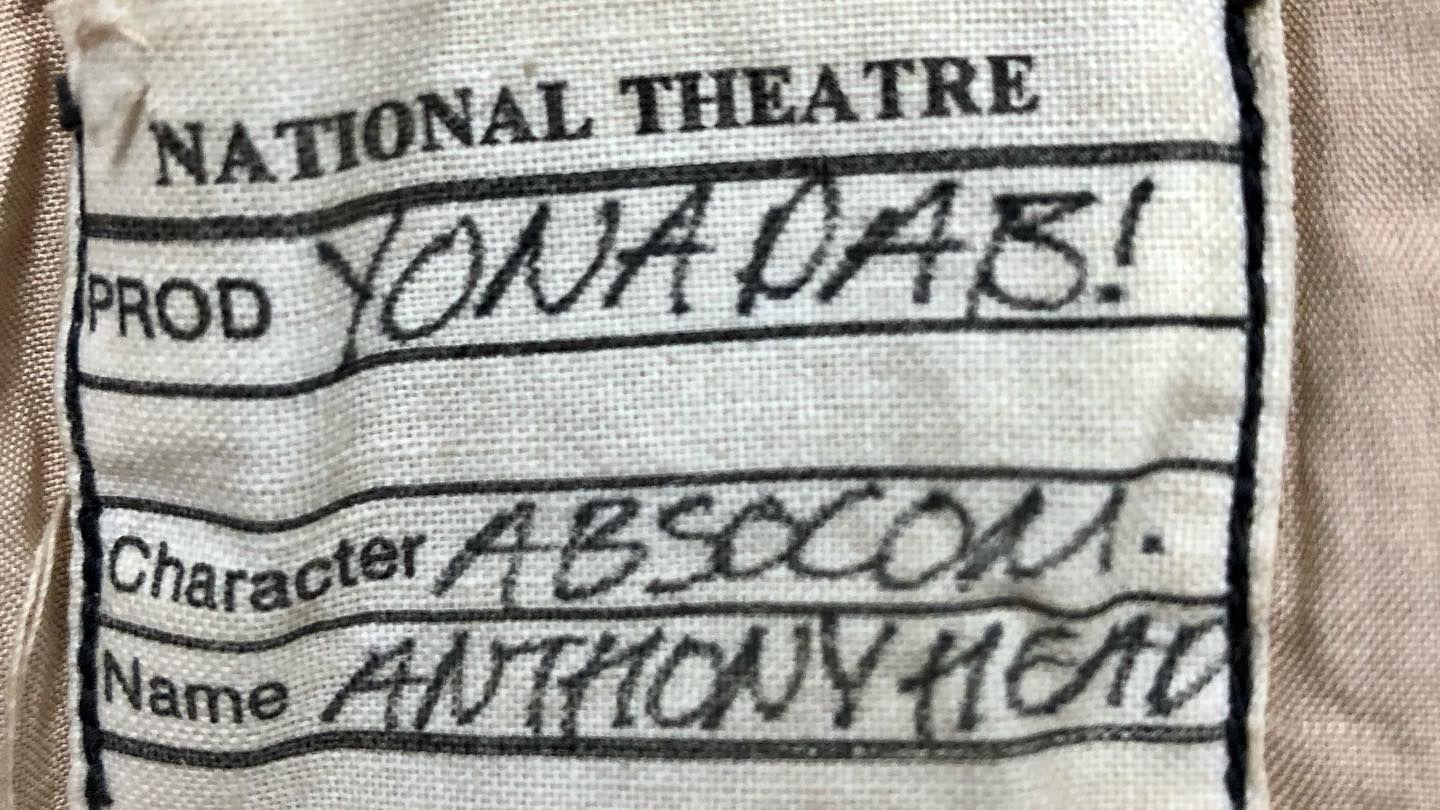 A white piece of fabric has been sewn onto the garment. It says National Theatre at the top, and has the name of the production, character and name of the actor written on it. 