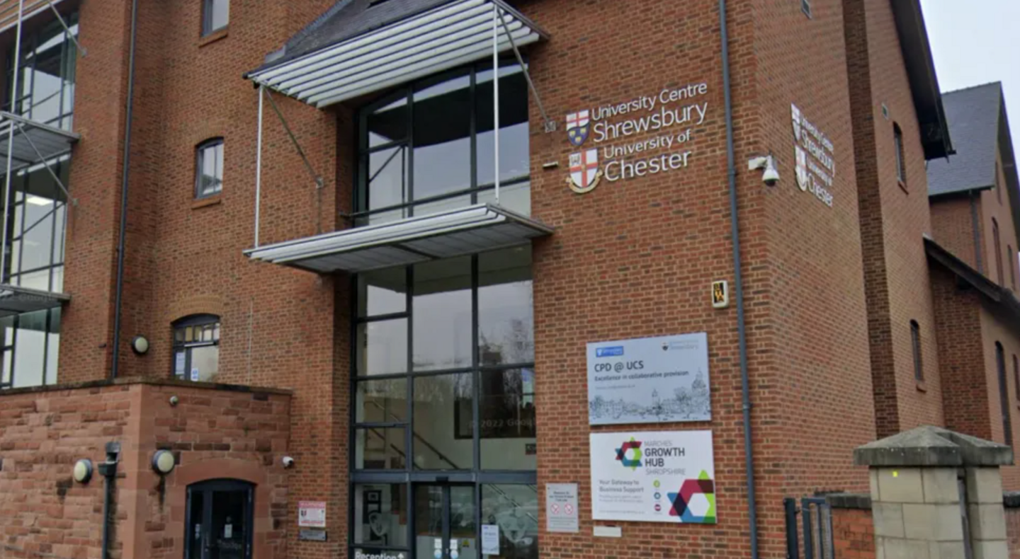 The main entrance to a red brick building with a large section of glass int he middle that spans over several floors