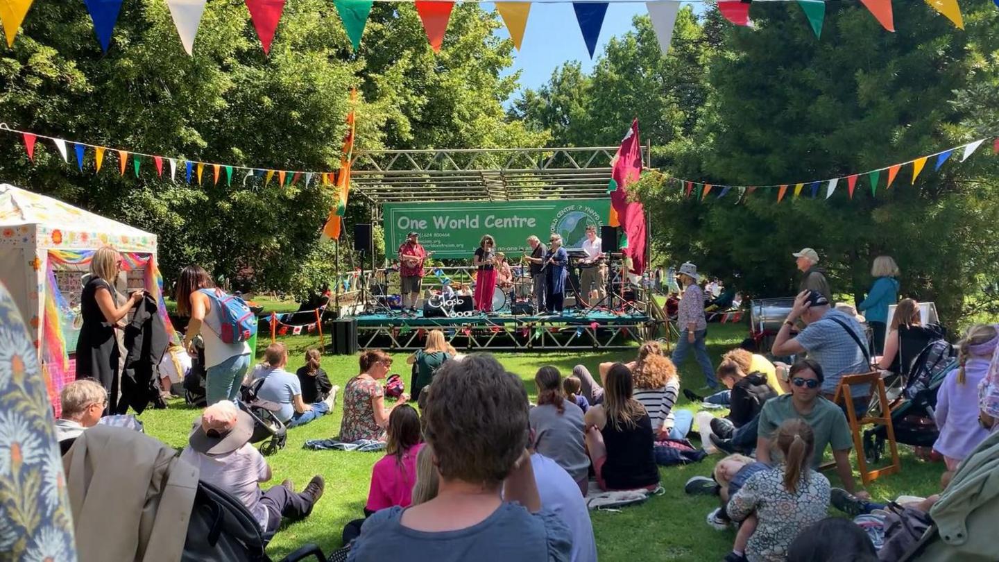 Crowds are sitting and standing around a One World Centre stage on the grass. A band is performing and there is colourful bunting all around.