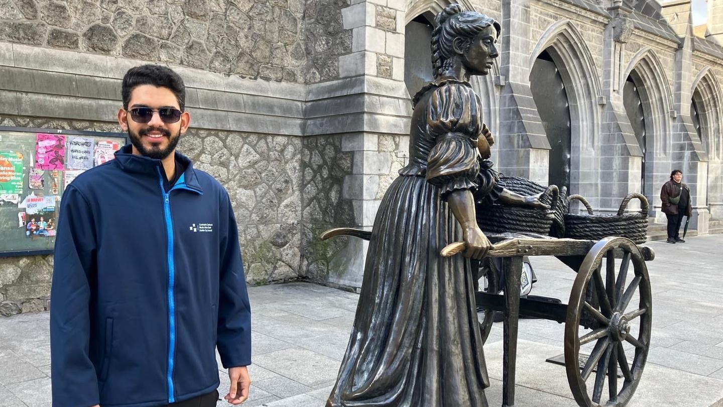 A man with sunglasses and a navy coat stands beside the famous statue of Molly Malone.