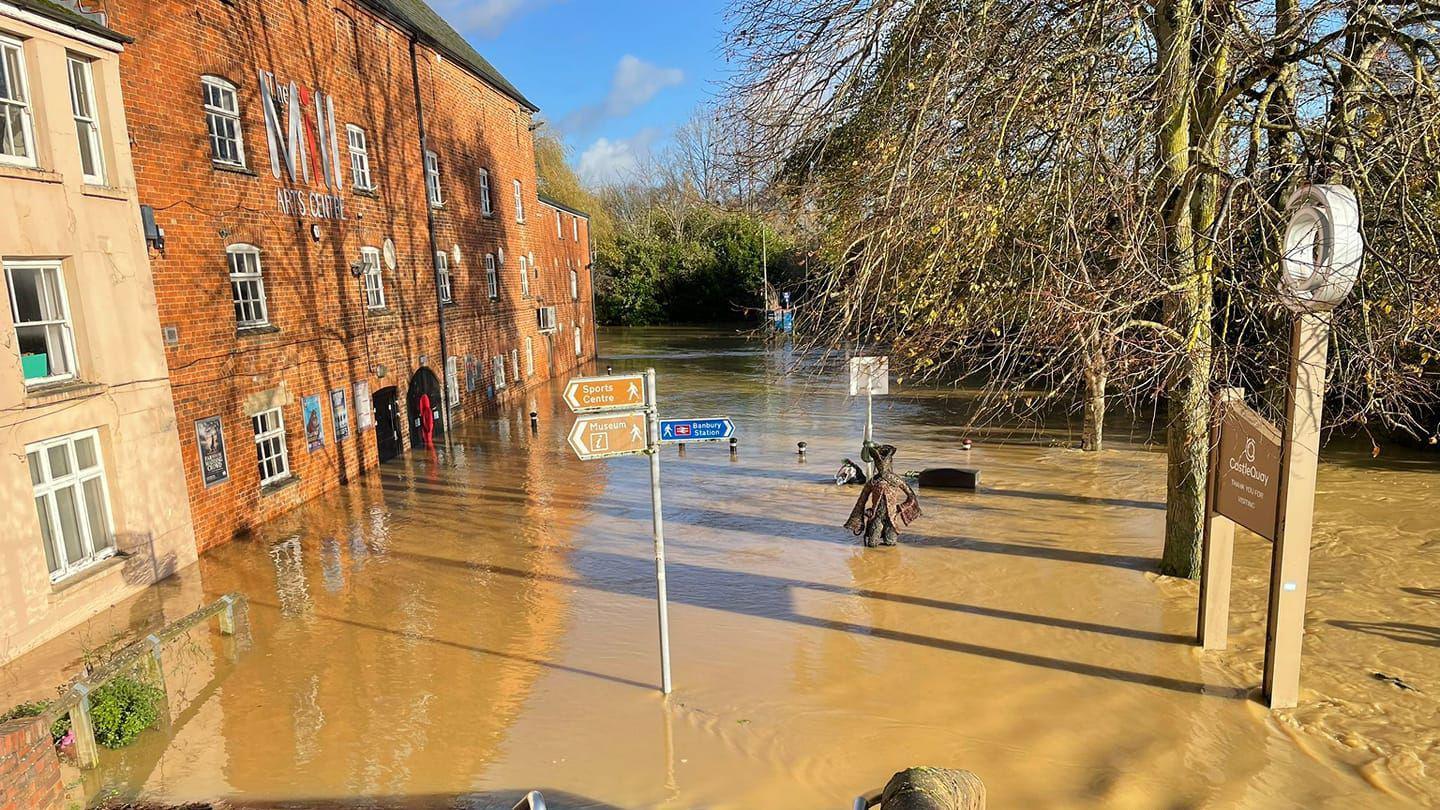 A red-brick building on the left looks like it is on the edge of a lake as it is surrounded by flood water.