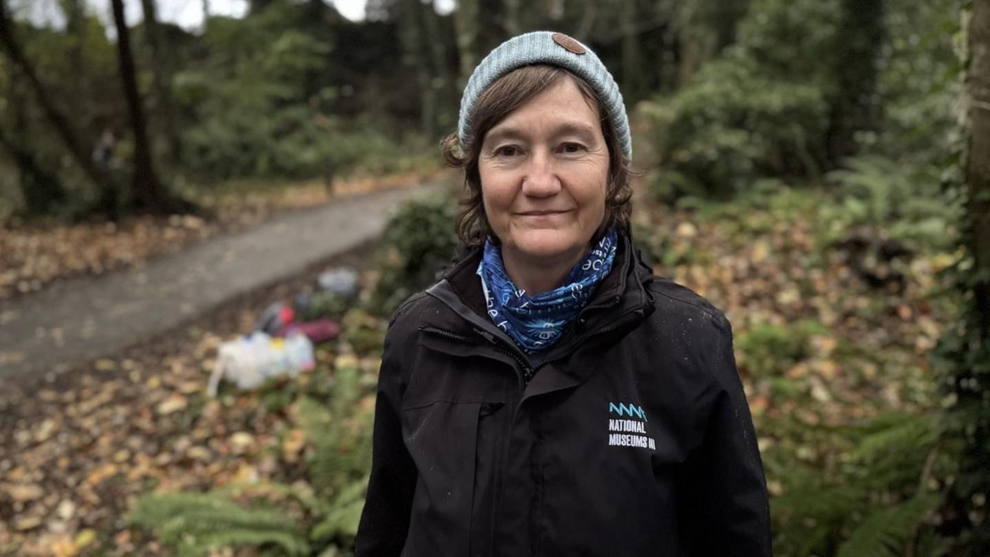 Helen James, a woman with dark hair, dark jacket, light blue hat and darker blue scarf. She is standing in a forest area.