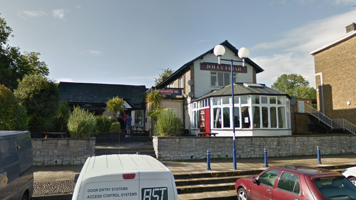 The Jolly Friar Pub sits on a slight elevation with steps leading to the entrance. It is surrounded by a small stone wall. It has a conservatory extension underneath a sign that says "Jolly Friar".