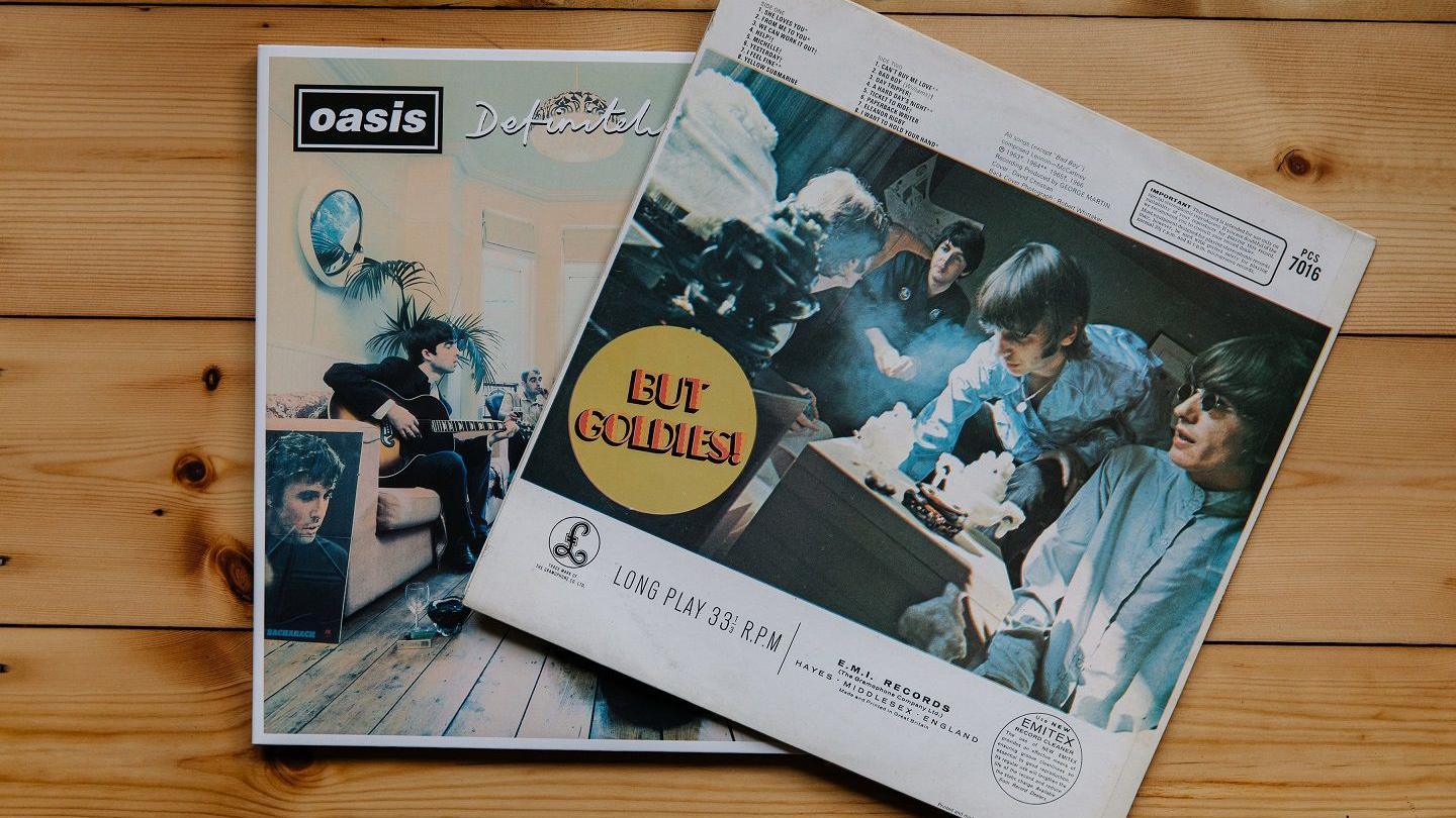 A vinyl album cover of The Beatles' compilation A Collection of Beatles Oldies partially covers a copy of the Definitely Maybe cover. The colour image on the Beatles' album shows the four members in a hotel room, with a vase and other objects on surfaces in front of them.