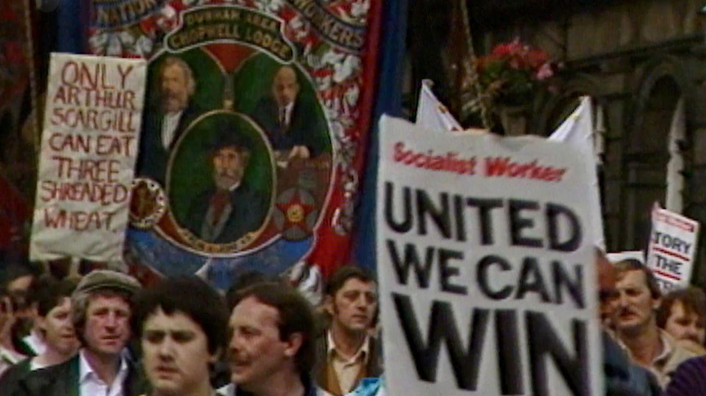 Men marching with banners, with one banner saying 'United we can win'. Another banner in the background reads 'Only Arthur Scargill can eat three shreaded wheat.' Another large, red and green banner is being held up which reads 'Chopwell Lodge' and has three painted portraits of men on it.