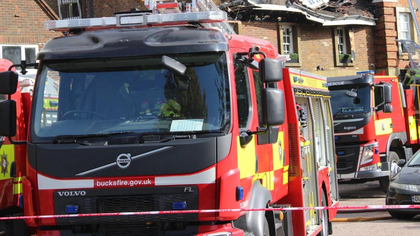 Red and yellow fire engine with bucksfire.gov.uk on the front in white lettering. There are engines behind and to the right of the one in the foreground. There is red and white emergency tape in the foreground. A building that has been damaged by fire is behind the engines.