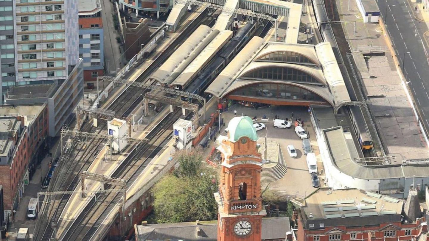 Railway tracks snake past Manchester Oxford Road train station, which is pictured from the sky. Cars can be seen park in the turning circle outside the station, with the view obstructed by the Kimpton Clocktower.