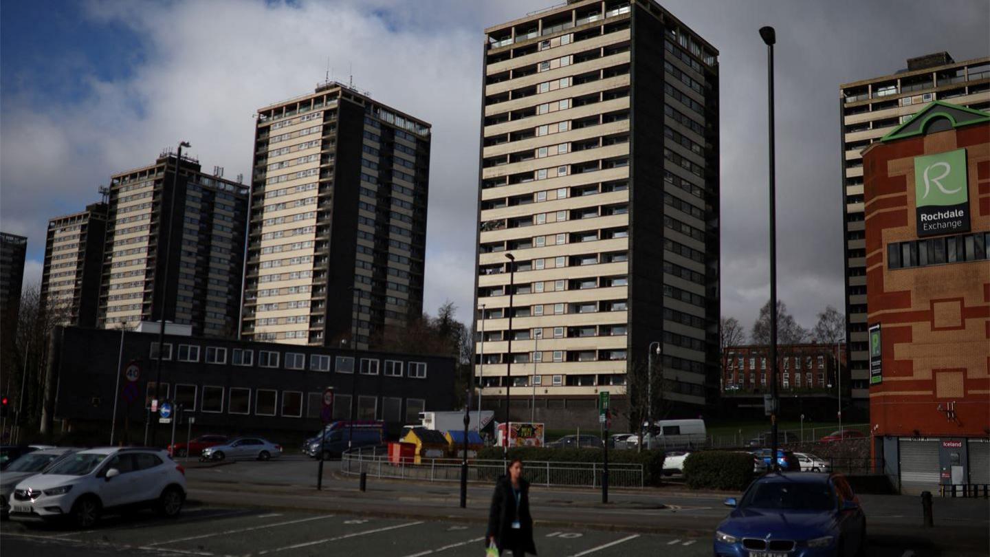 A woman walks in front of the Seven Sisters residential tower blocks in a car  park on a cloudy day in Rochdale