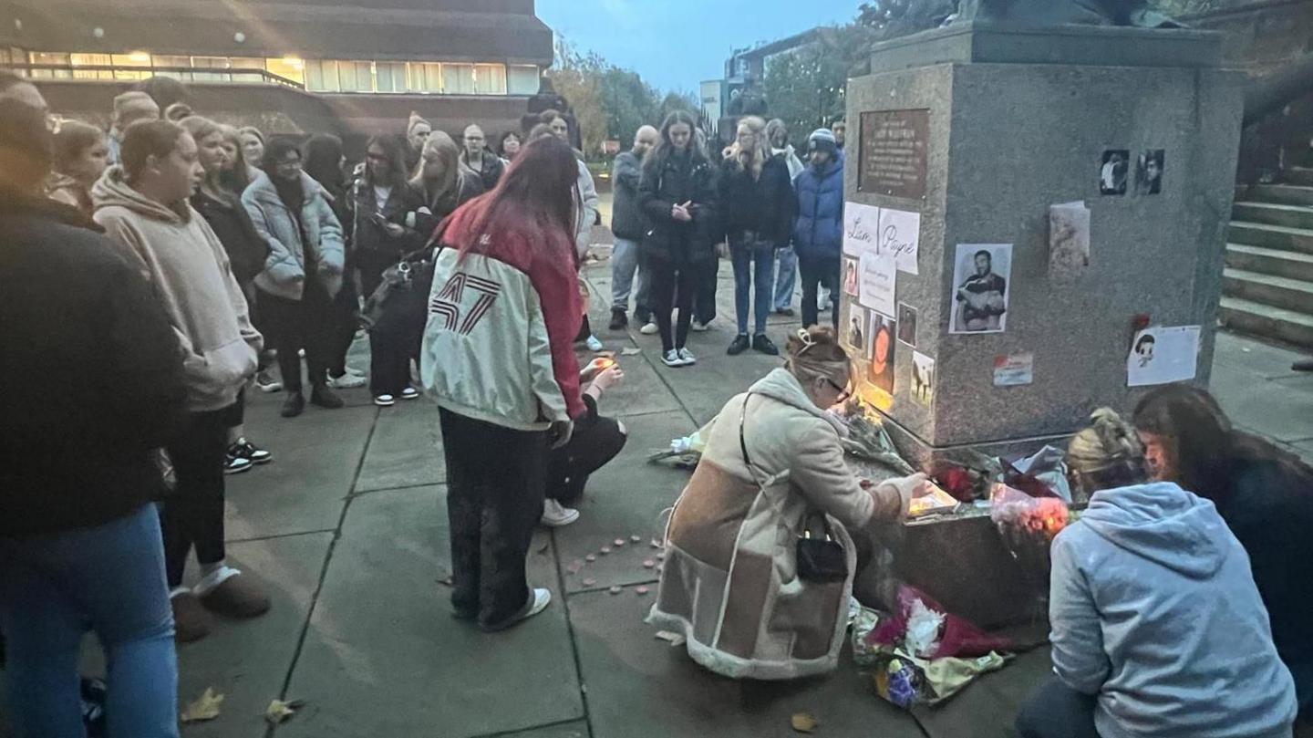 A line of people watch on as others light candles and kneel down at the foot of the statue where the tributes to Liam Payne have been laid.