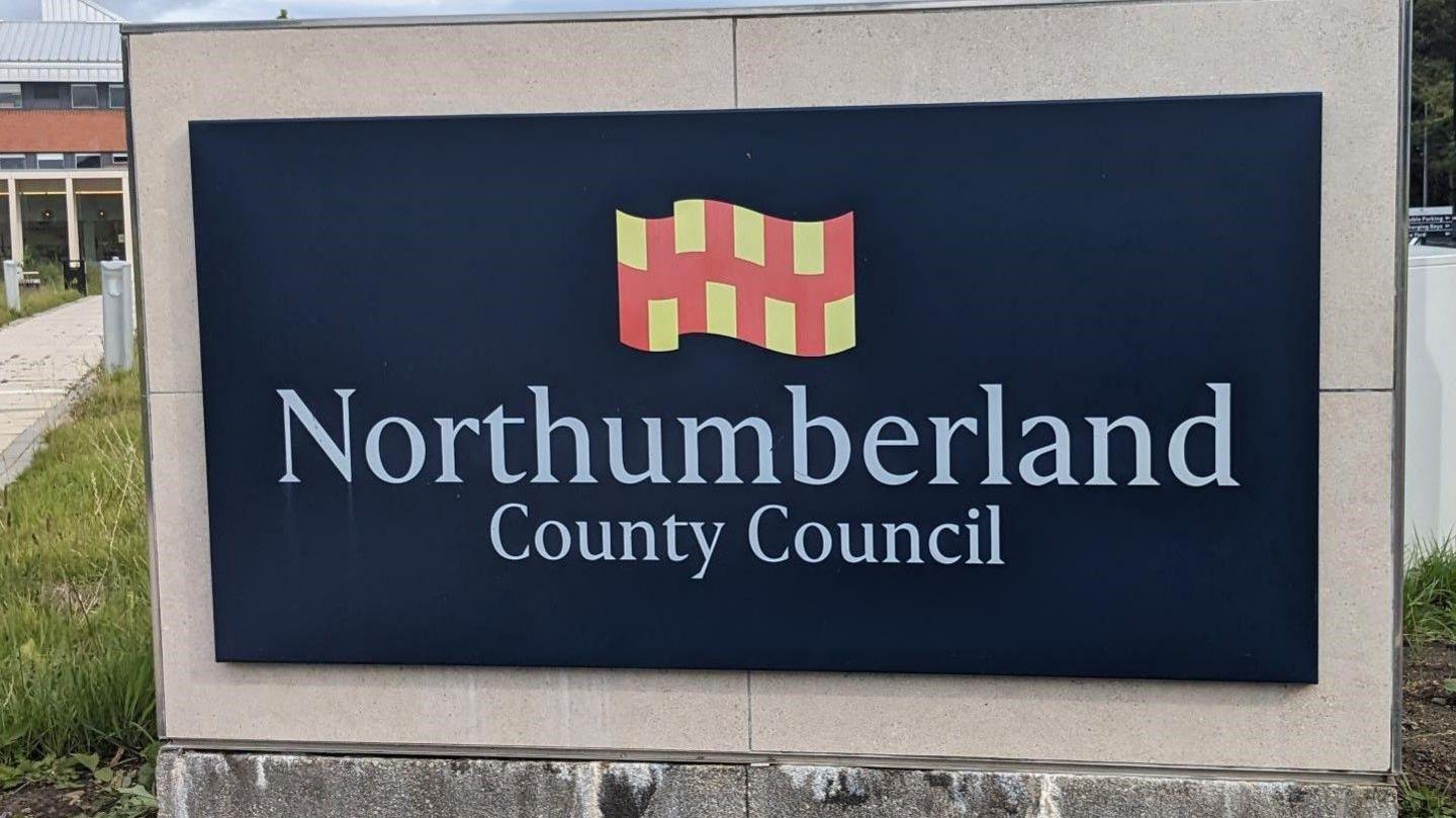 A navy blue sign in front of a building which reads 'Northumberland County Council'. The Northumberland flag made up of red and yellow squares is above the lettering.