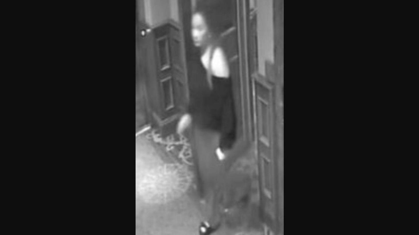 Black and white CCTV image of woman walking through door - she has long dark hair and is wearing a jumper of her shoulder and trousers.