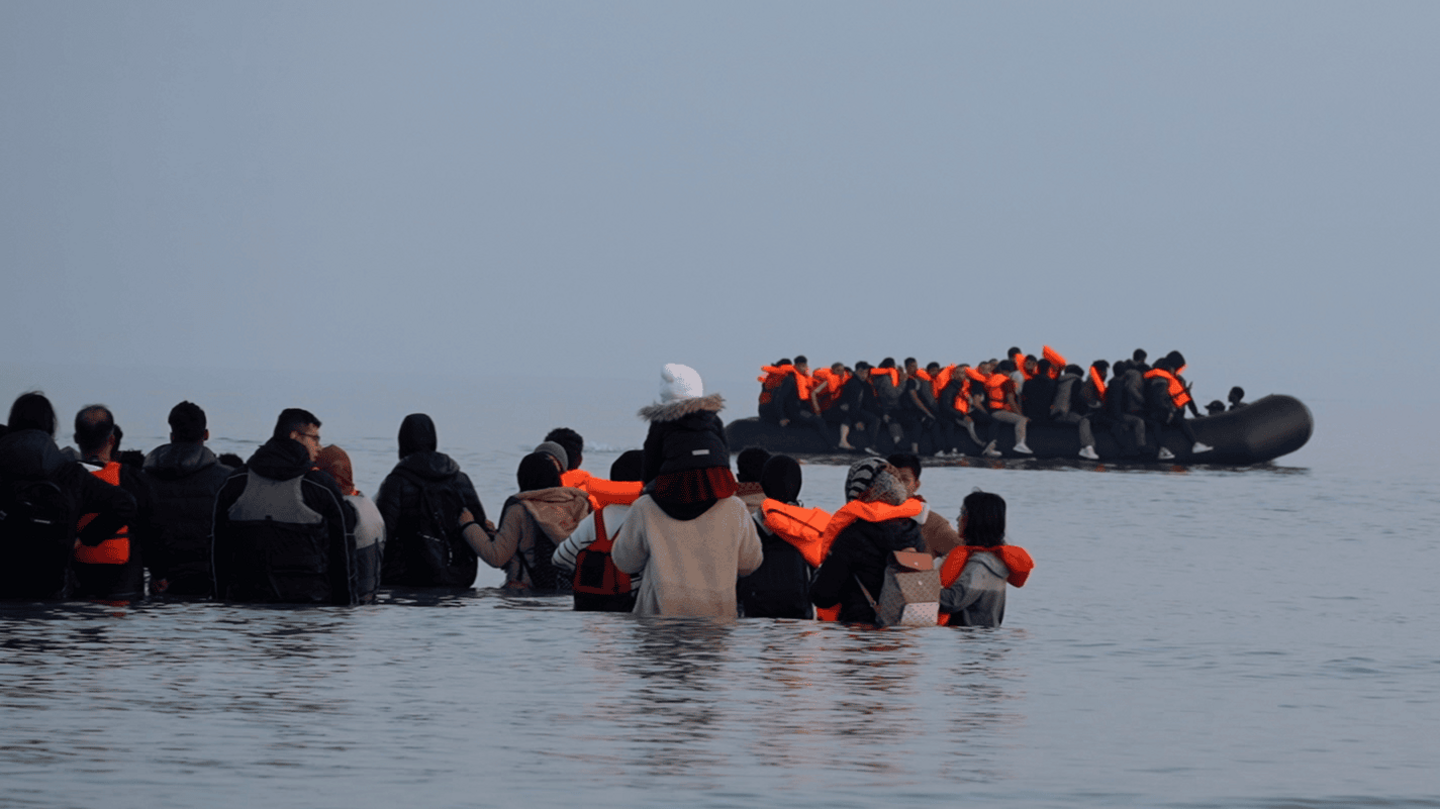 A group of people stand in waist-deep water with an overloaded inflatable boat in front of them