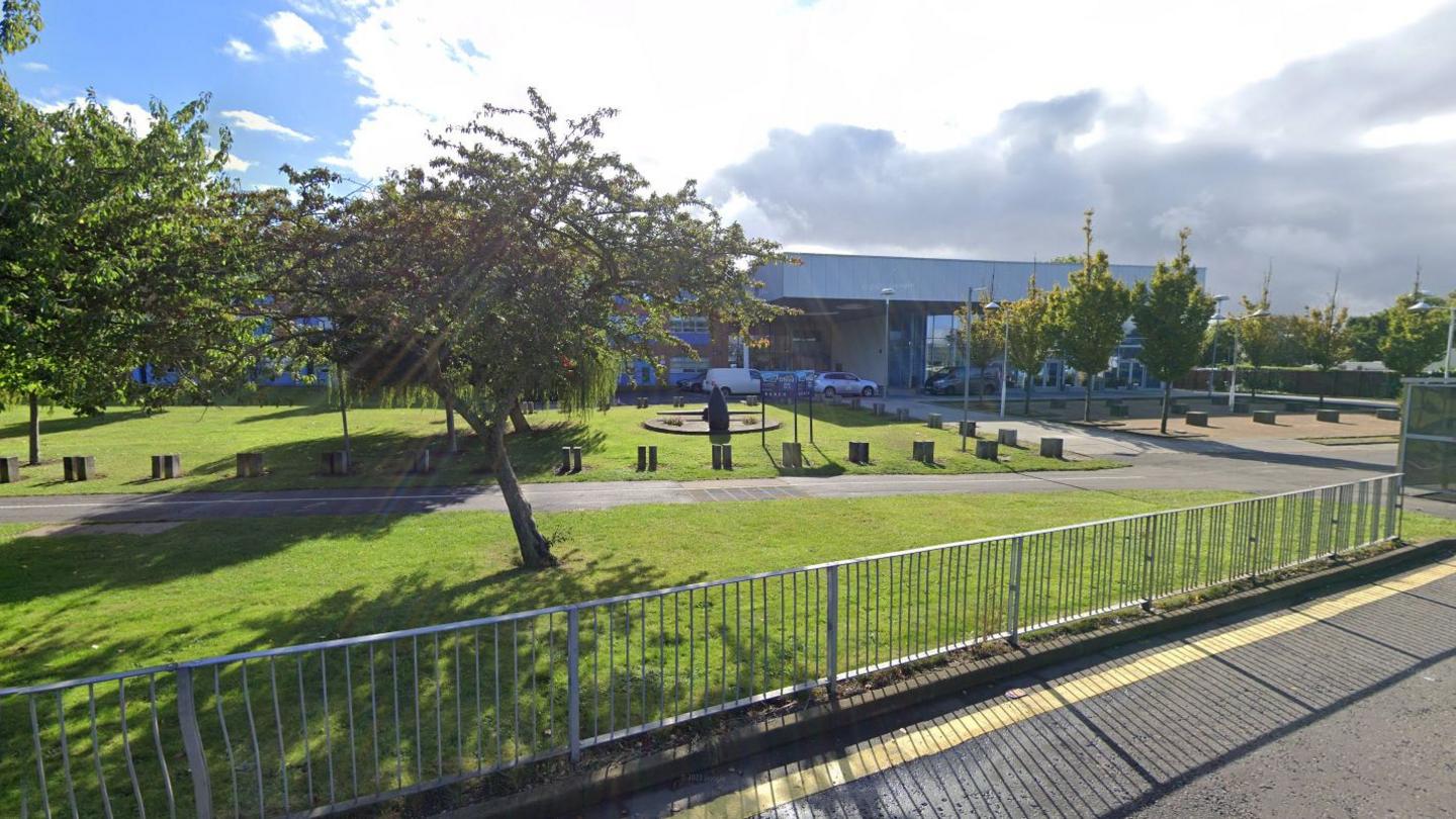 The outside of a school, which is a modern two-storey building, with railings, grass and trees at the front.