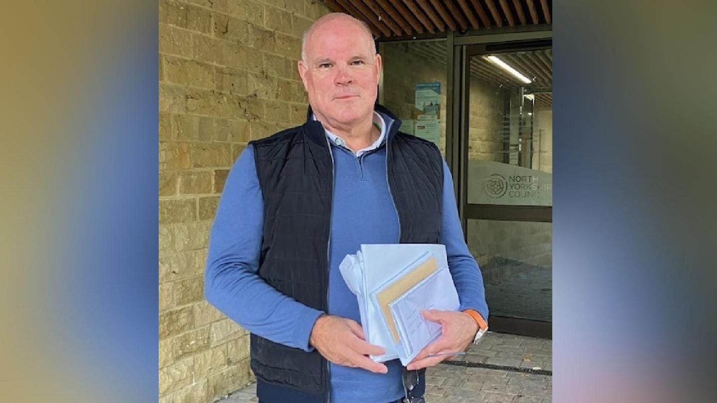A bald man wearing a light blue sweater and a dark blue gilet. He is holding a large white paper envelope and a bunch of forms and letters between his two hands.