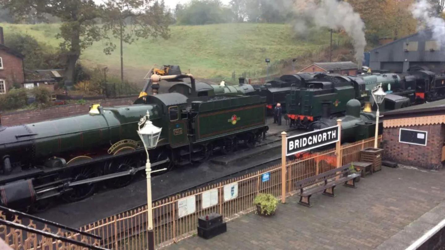 Two green steam trains with steam coming from their funnels at a station platform with metal railings, plants and a sign which says Bridgnorth