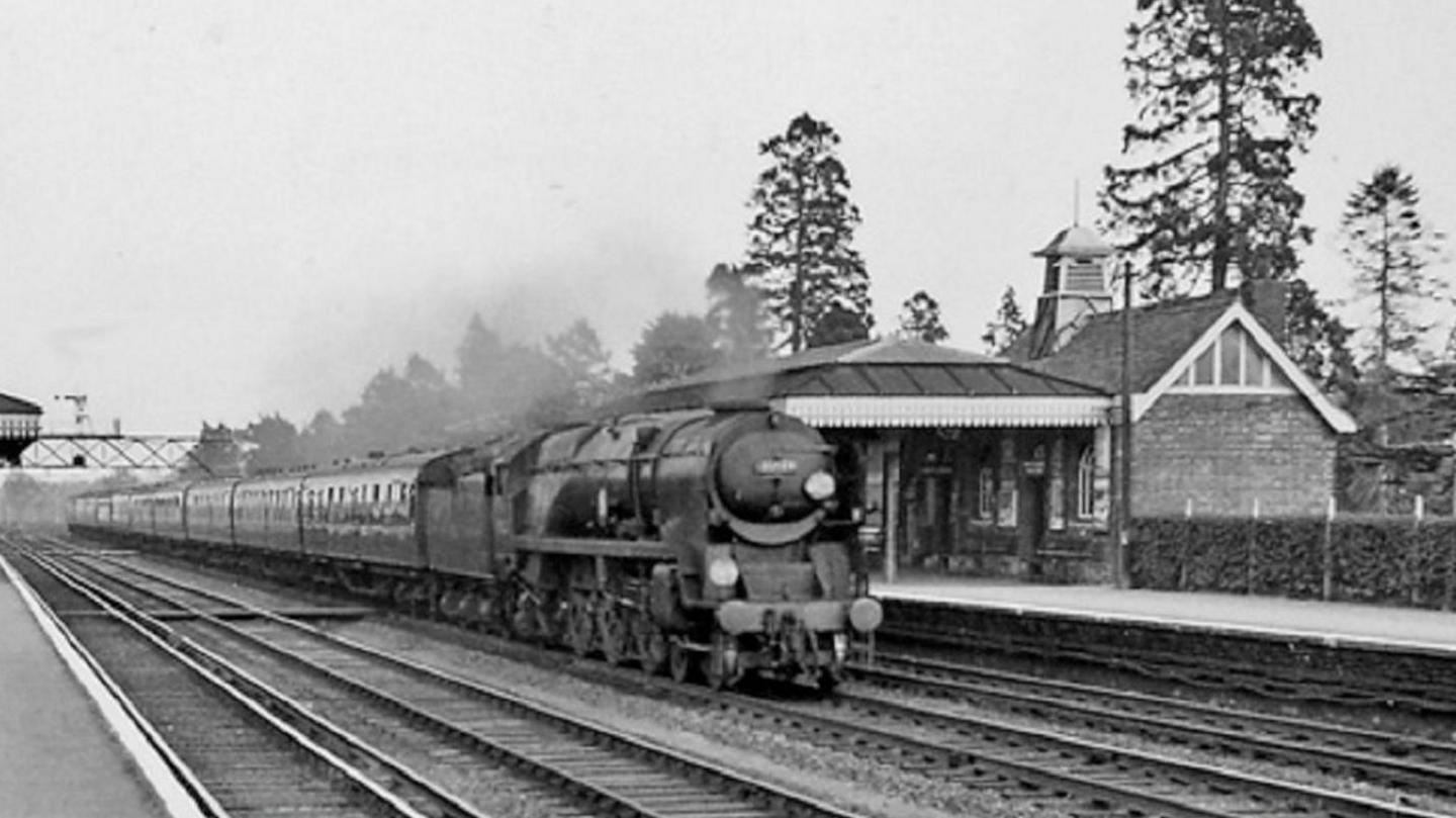 A black and white image of a train at a railway station. The train has smoke coming from the top of it.