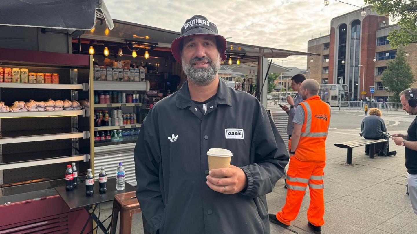 A man in a black jacket and black bucket hat stands with a coffee in front of a street food van, with shelves of drinks and pastries behind him.