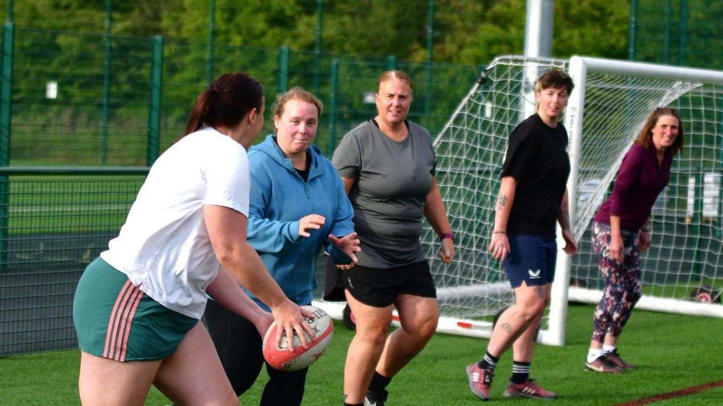 A line of five women playing rugby. To the left, the player closest to the camera is about to pass the ball. The second player is waiting to catch it. The women are wearing shorts and t-shirts of various styles and colours.