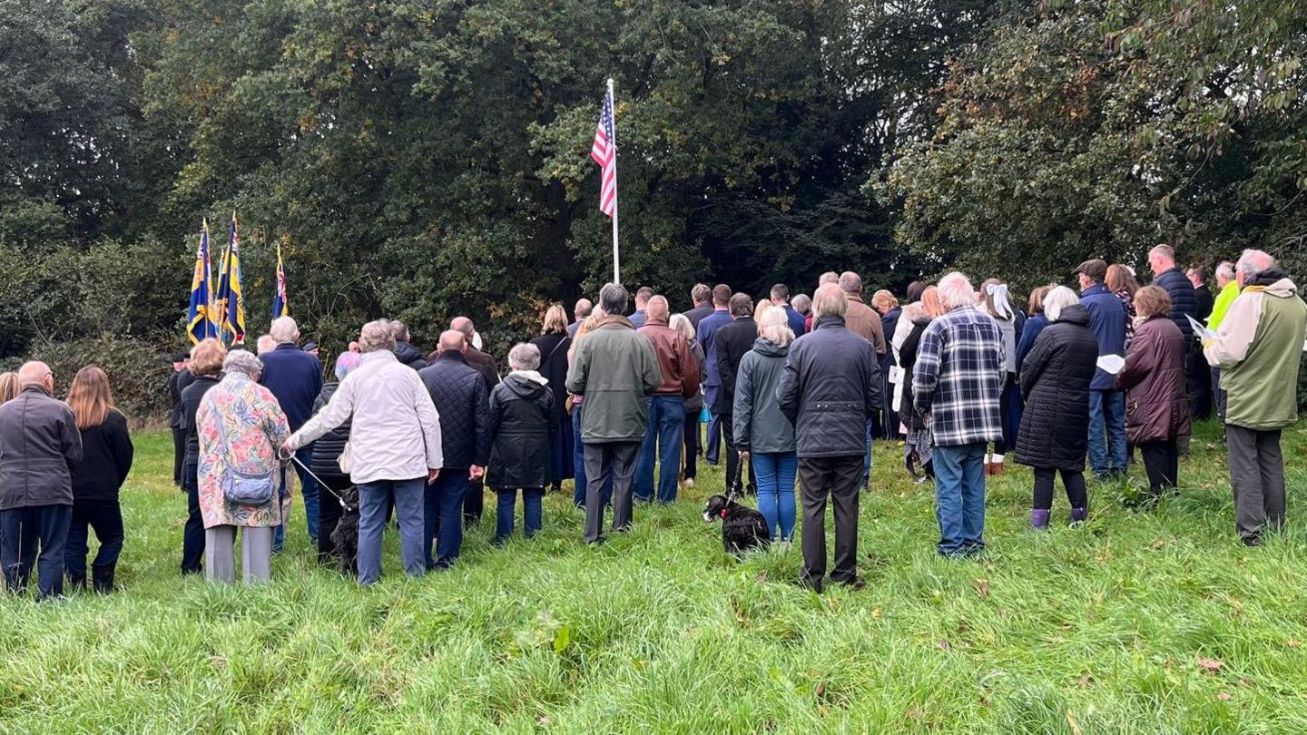 A crowd seen from behind standing around an outdoor flagpole with the USA flag. There are trees and a field.