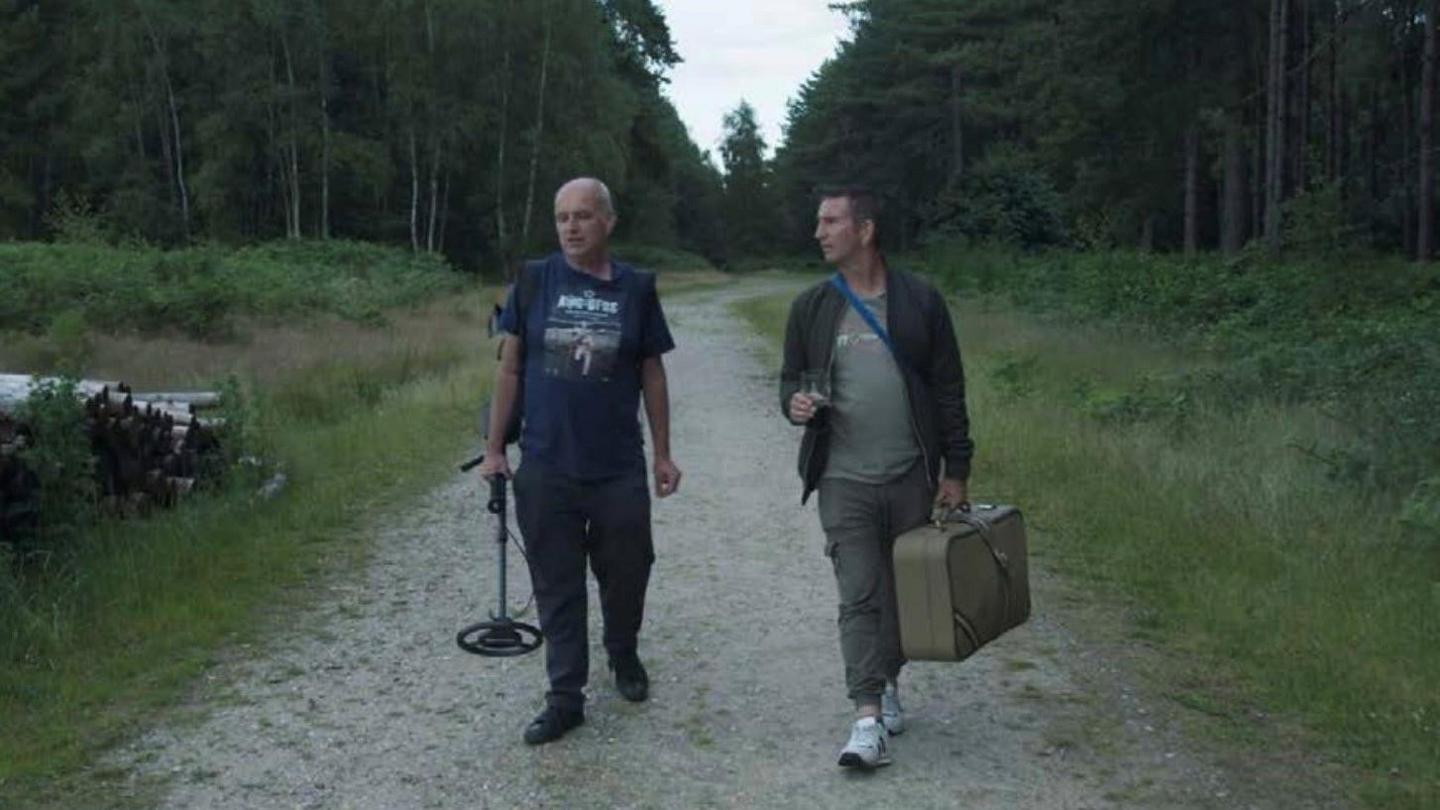 Two men, one on the left holding a metal detector and the one on the right with a square shaped bag, are walking down a pathway in the middle of a forest.