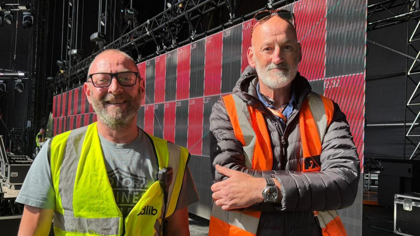 Marc and Andy wearing hi-vis jackets on a stage. They are both smiling at the camera. Both have glasses and grey beards.