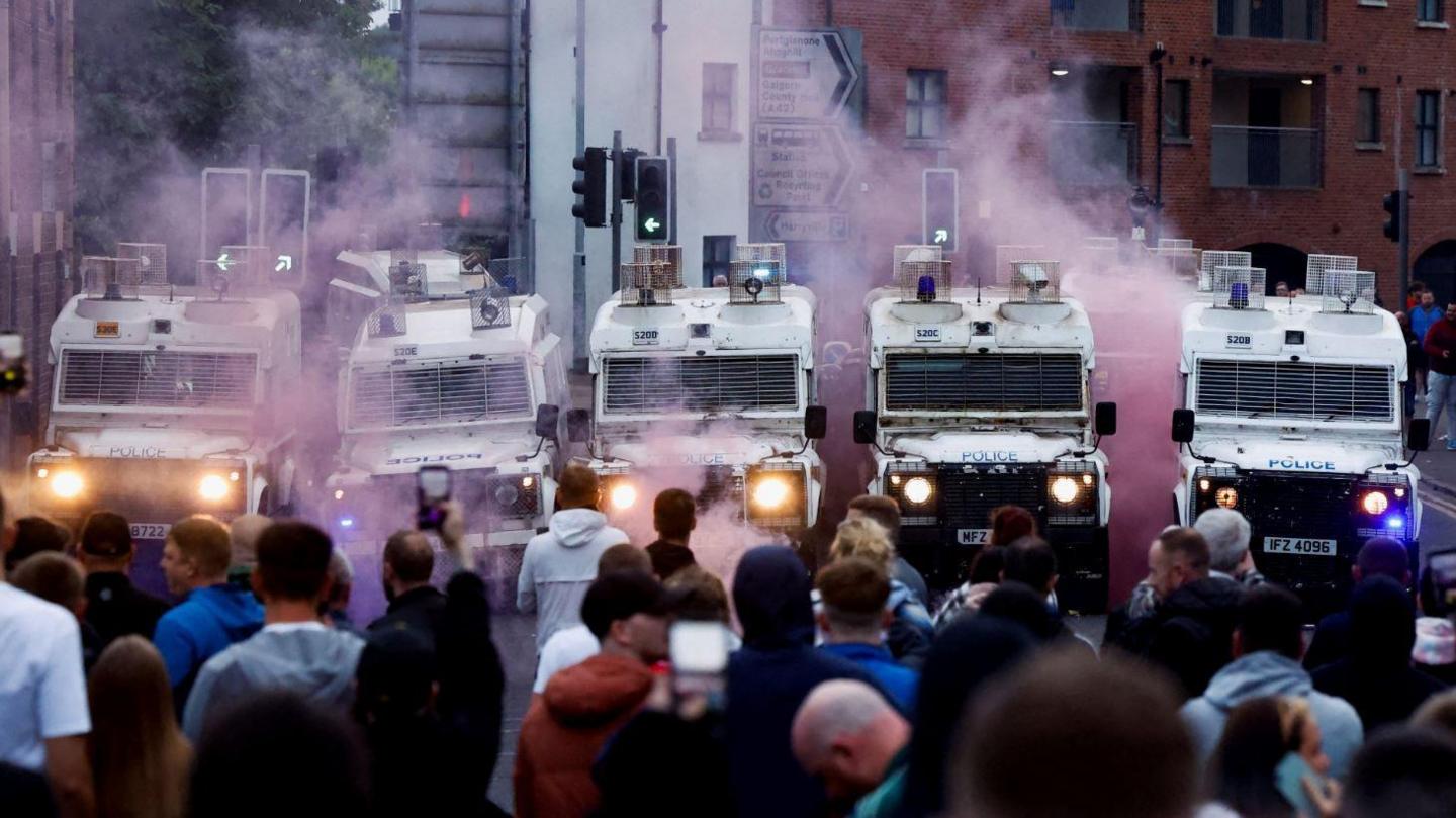 A crowd of people gathered in front of five white PSNI Land Rover vehicles. Pink smoke is billowing from the scene. Some of the people are holding up mobile phones to film the Land Rovers. Behind the line of police vehicles are a set of traffic lights and a street sign as well as a modern red-brick apartment block.