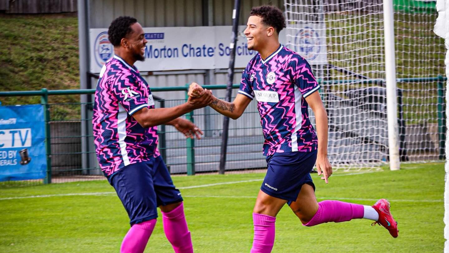 Two players celebrate a goal with a fist bump, as they run together. They are to the left of the goal and wear the pink and blue kit as described above.