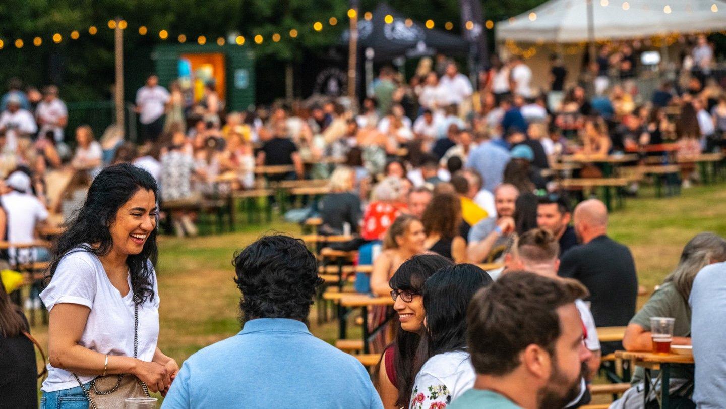 Warwick Castle Dining Club event during a previous tour. People are sitting at tables set up on a grassy space outside with food and drink. There are strings of lights in the background and the canopy of a stall.