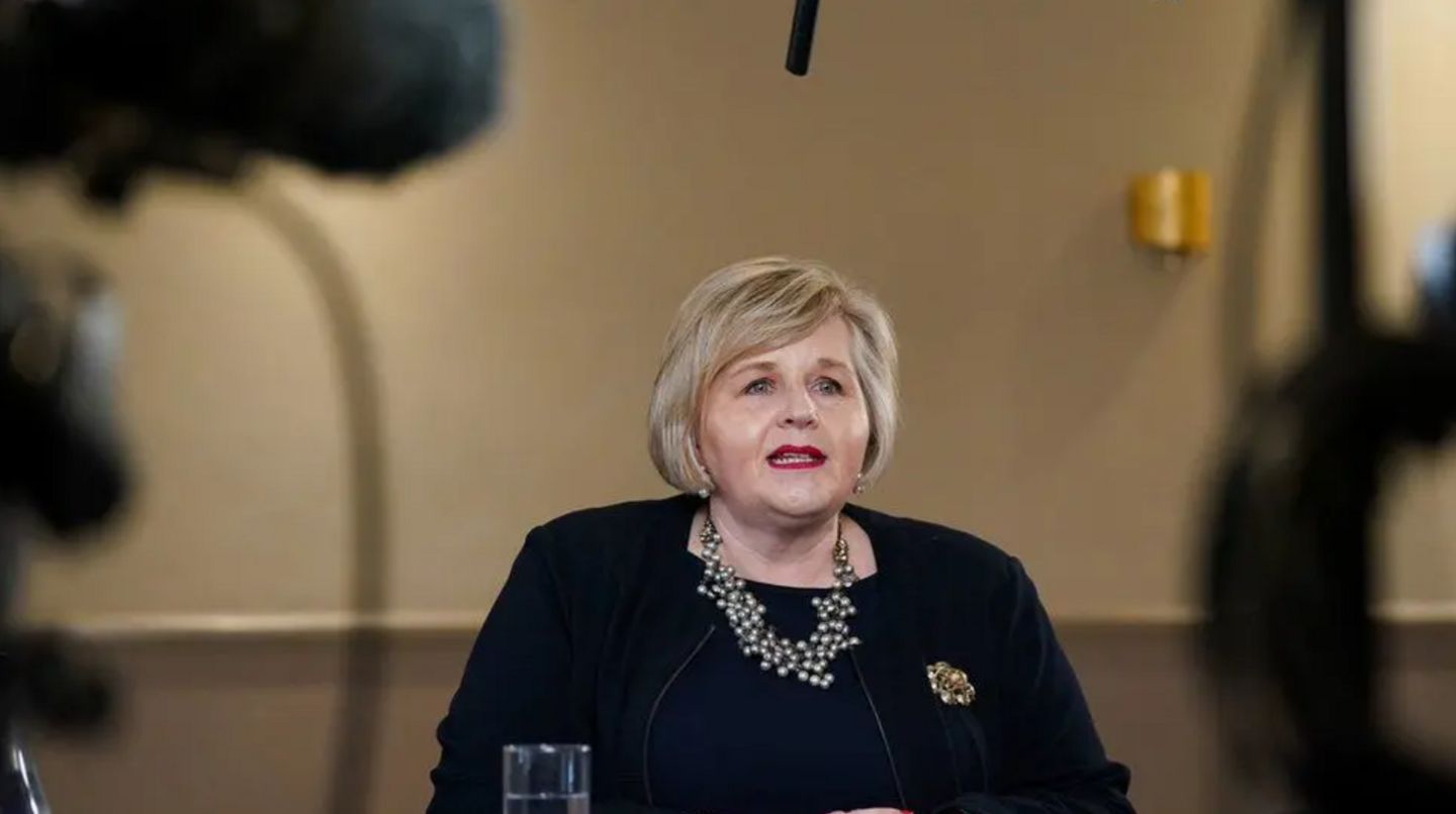 Donna Ockenden seated at a desk at a press conference. She is wearing all black and has a large necklace on. The background is slightly blurred but cameras can be seen. 