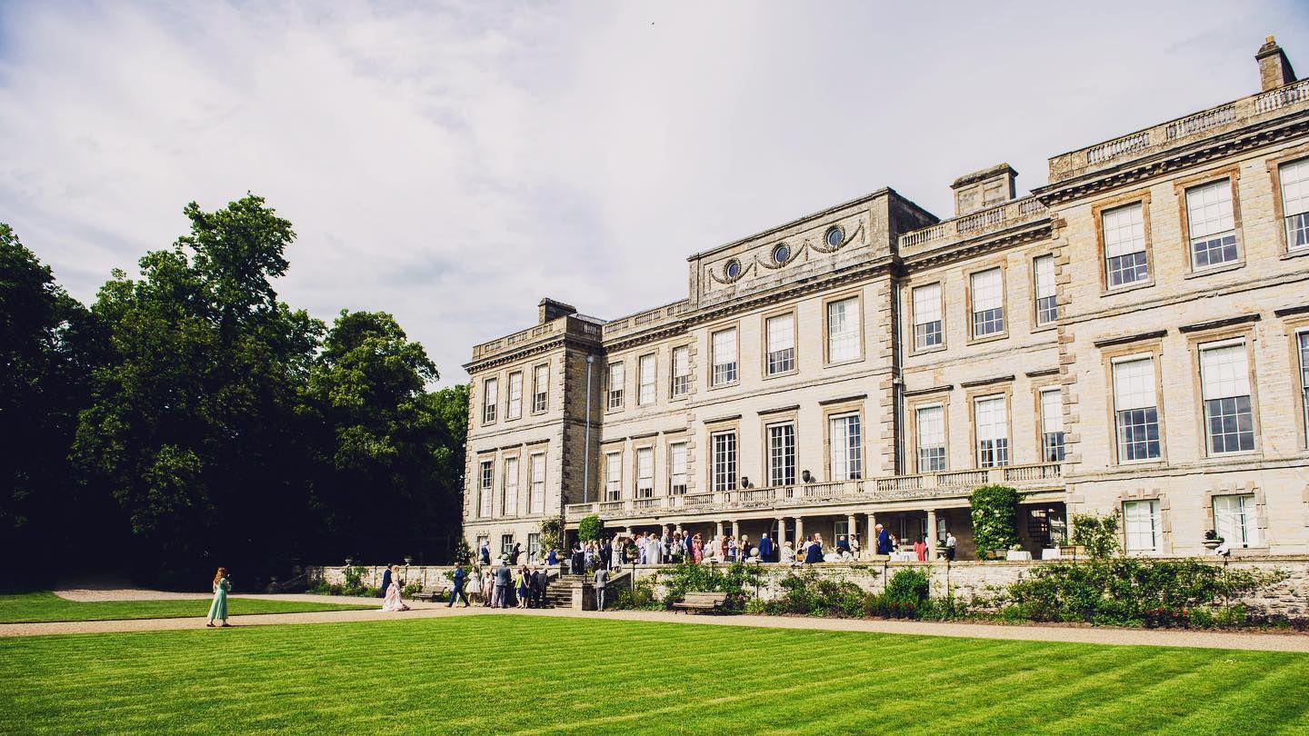 Ragley Hall, a large Robert Hooke Palladian mansion with party goers on the front steps and a grass lawn in front of the building. 