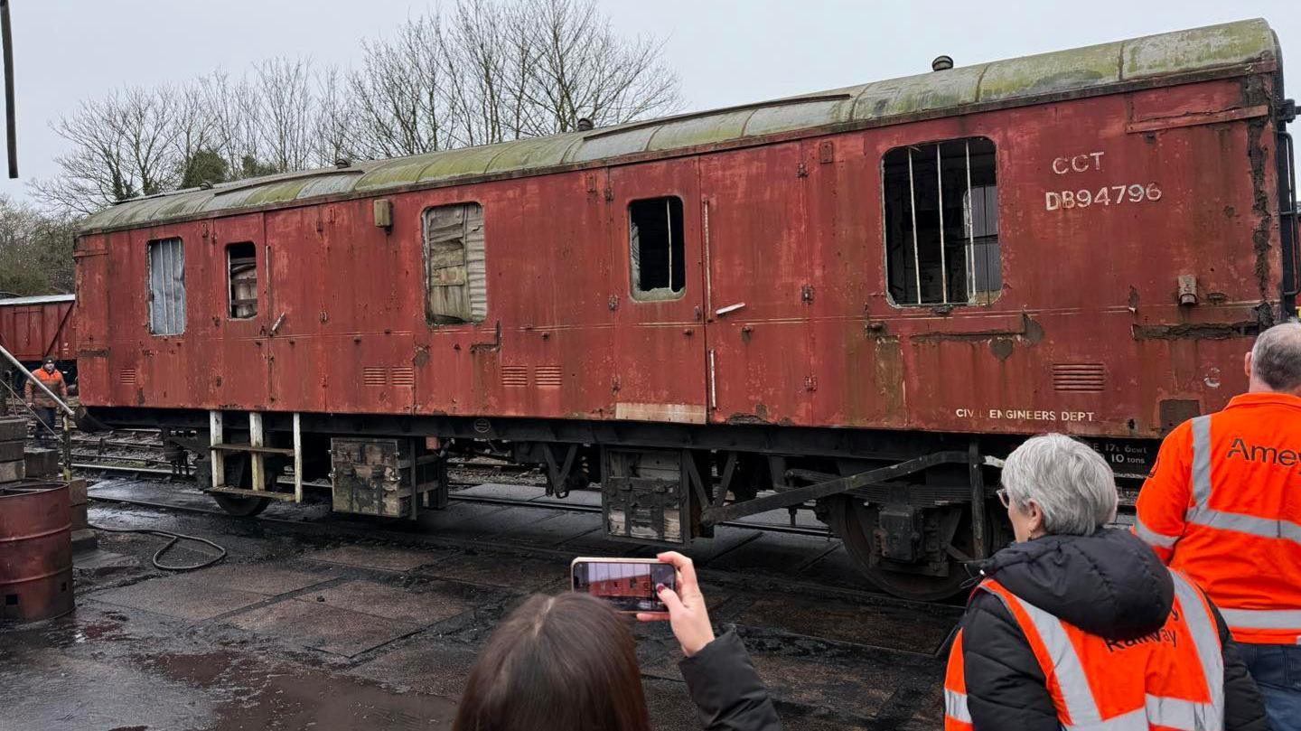 A derelict red station wagon parked on the platform with members of the Amey firm standing in front of it- wearing orange and silver high-visibility jackets.