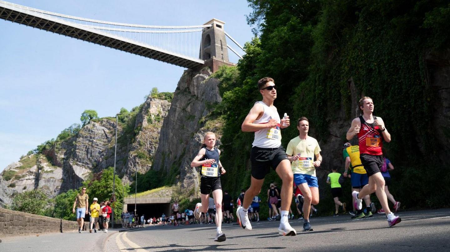 People in running gear are running on a road. In the background we can see the Clifton Suspension Bridge.
