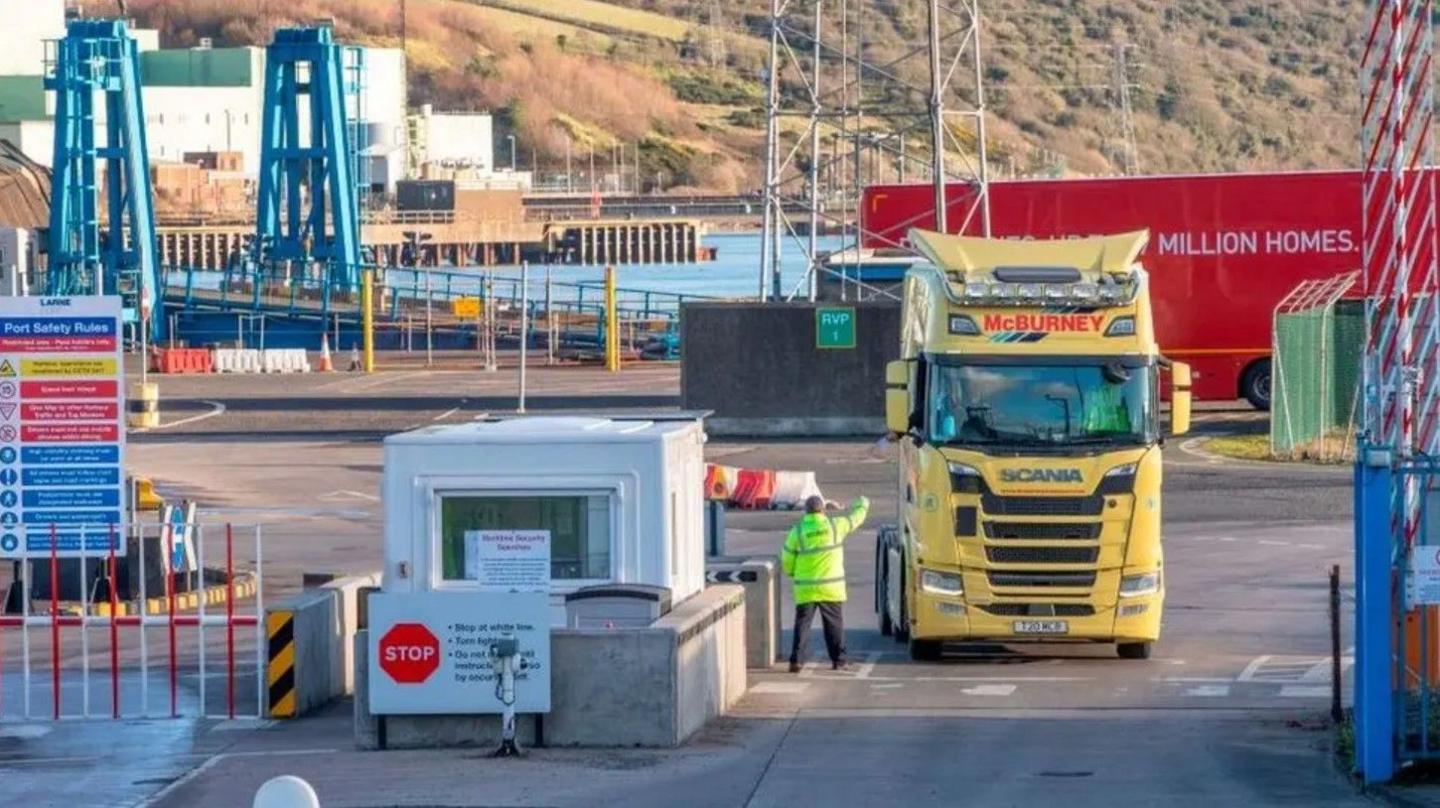 Yellow lorry at a port being directed by a man in a high-vis jacket.