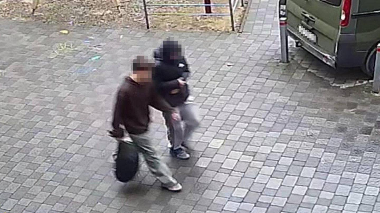 A still from a security camera looking down on a side view of two older teenage boys  in casual clothes walking along a paved street. Their faces are blurred. The one nearest the camera is holding a black rucksack in his right hand.