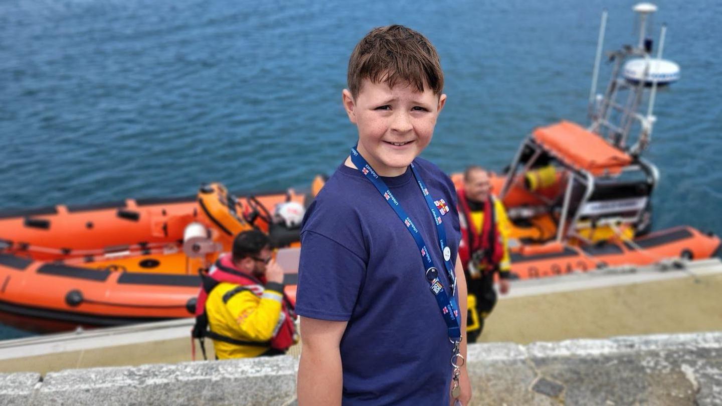 A young boy with brown hair wearing a blue shirt with the RNLI logo on it. In the background are two men wearing life jackets and an orange lifeboat on the water. 