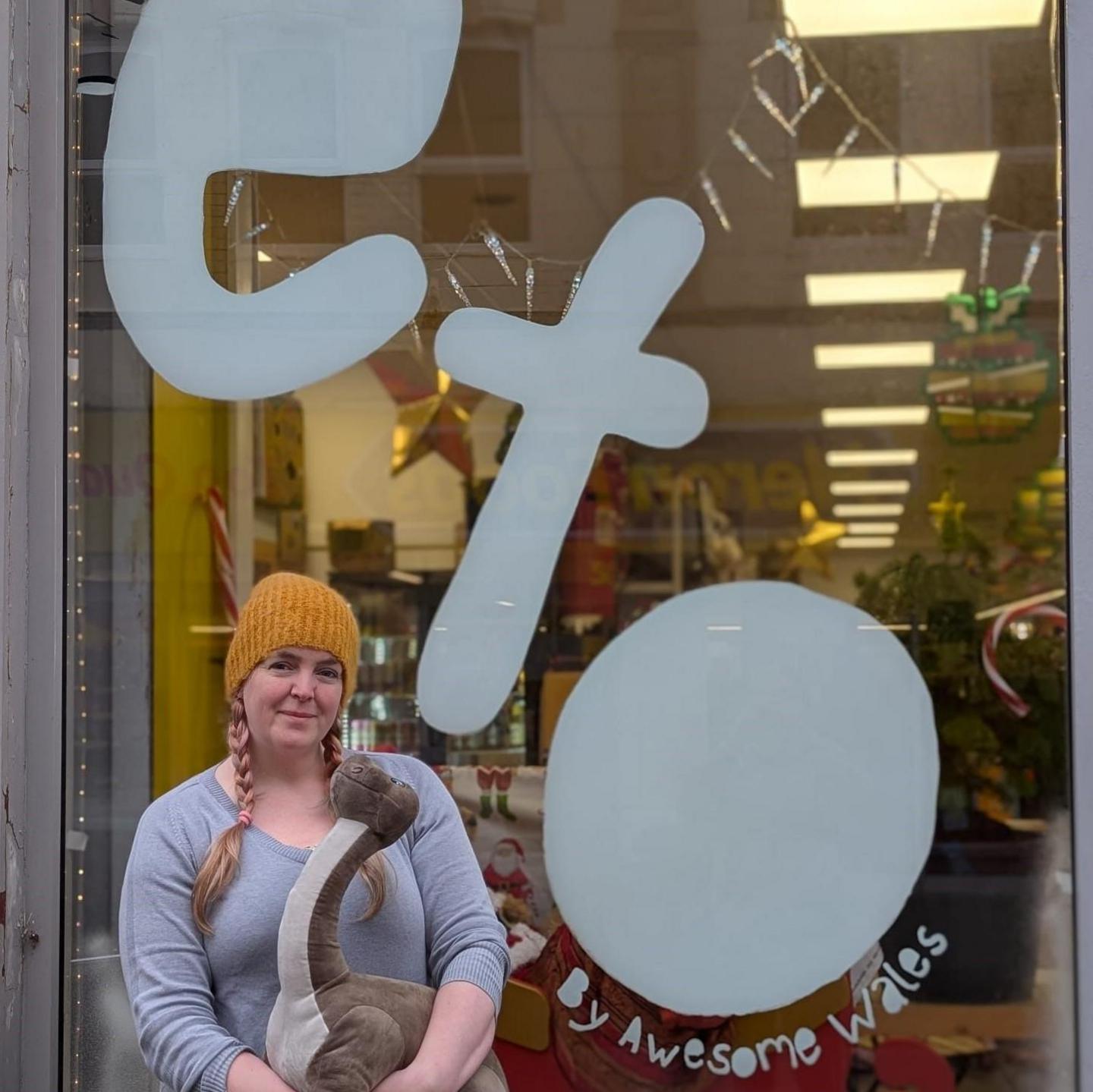 Amy Greenfield stands outside of her store in Barry, holding a dinosaur stuffed toy in her arms. She wears a mustard woolly hat and a grey jumper and has pink hues in her ginger plaited hair. She smiles into the camera. 