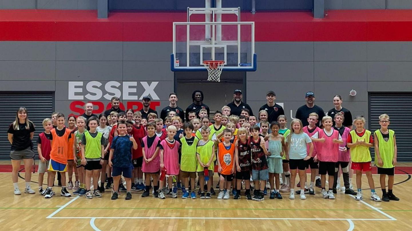 Children, university staff and police officers stand together as a group for a picture on the basketball court. They are all looking at the camera and are stood beneath a basketball net.