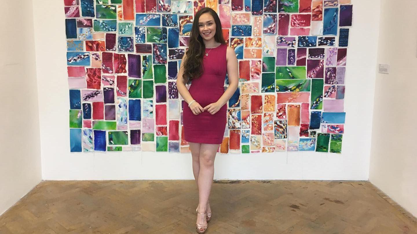 A woman wearing a burgundy dress and a pair of high heels. She is stood in front of a background comprised of rectangular shapes featuring a number of different colours.