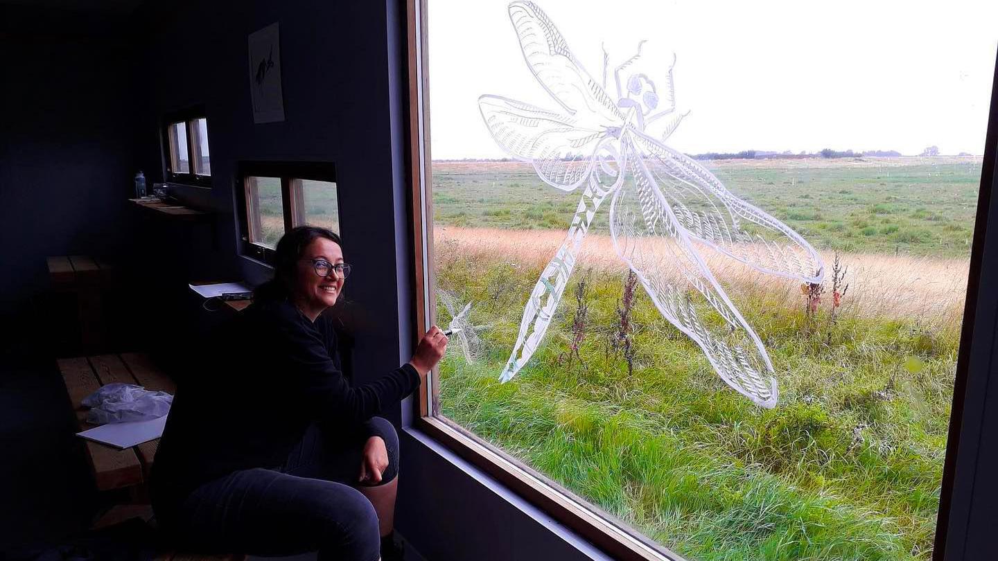 A woman is sitting on a bench in a building which has a few very small windows and one very large window. On the large window the woman is painting a big dragonfly in what appears to be white paint. Grass fields can be seen outside. The woman is smiling and looking at the camera.