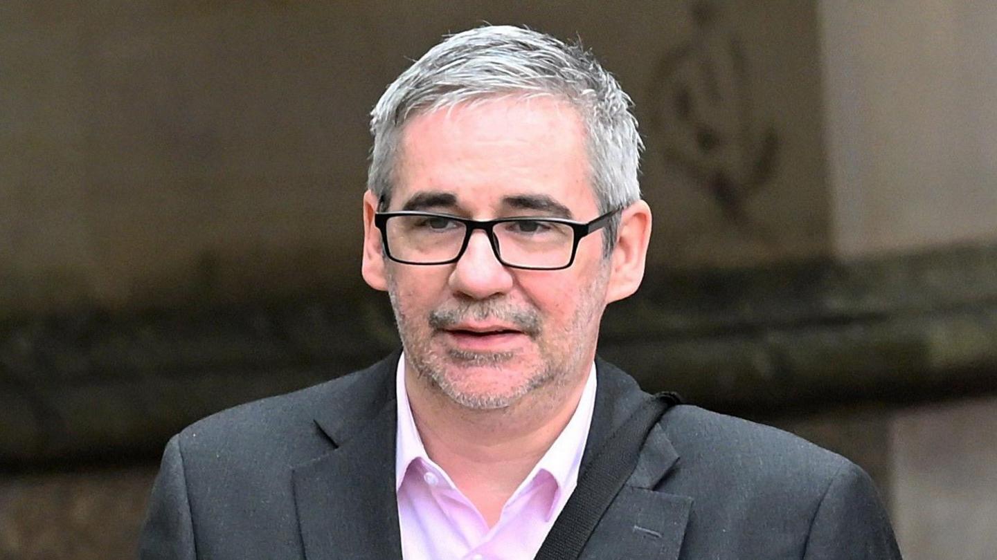 Iain Robertson wears a grey suit and pink shirt. he has short grey hair and wears dark glasses. He is walking outside a building