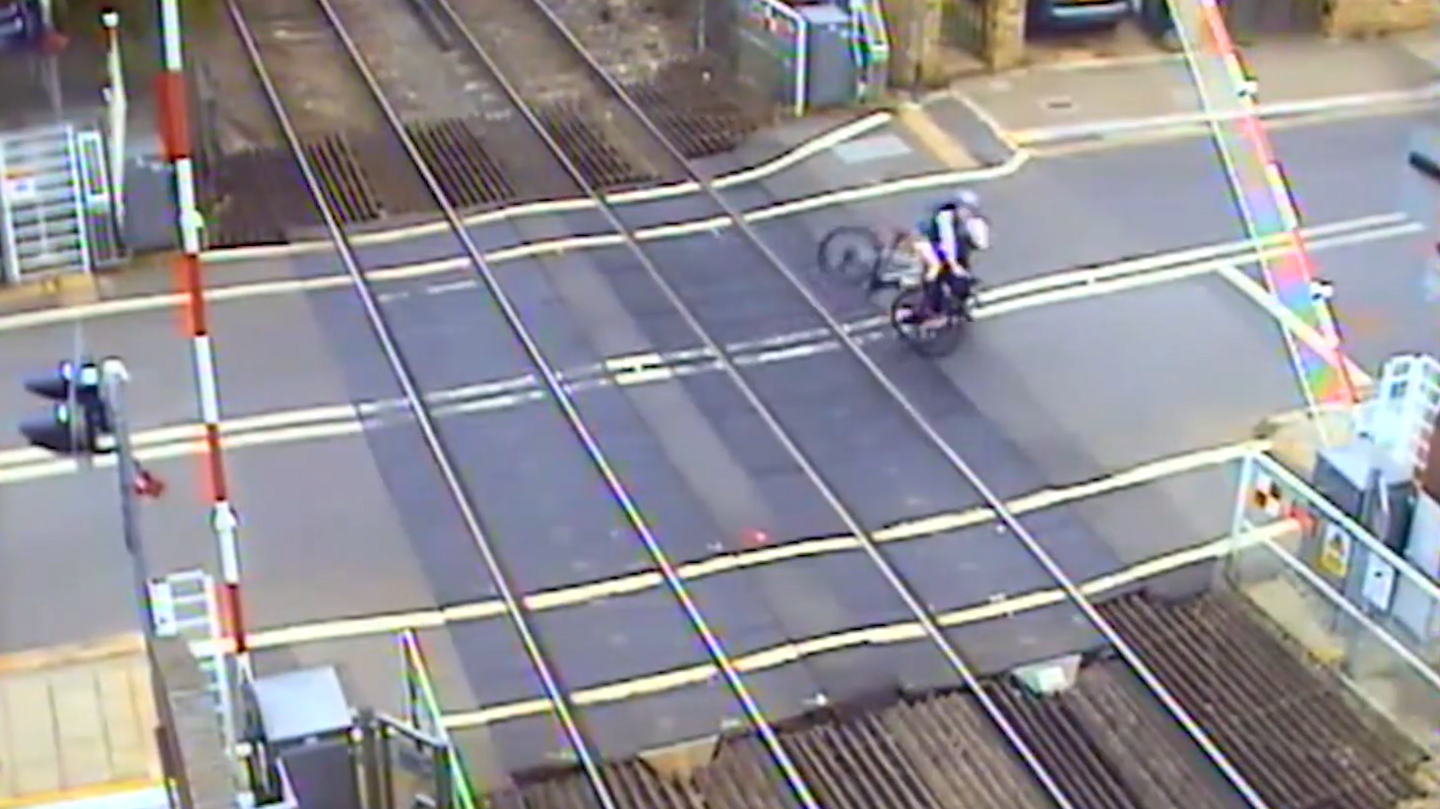 Two cyclists are falling next to each other on a level crossing, close to the track and a lowering barrier