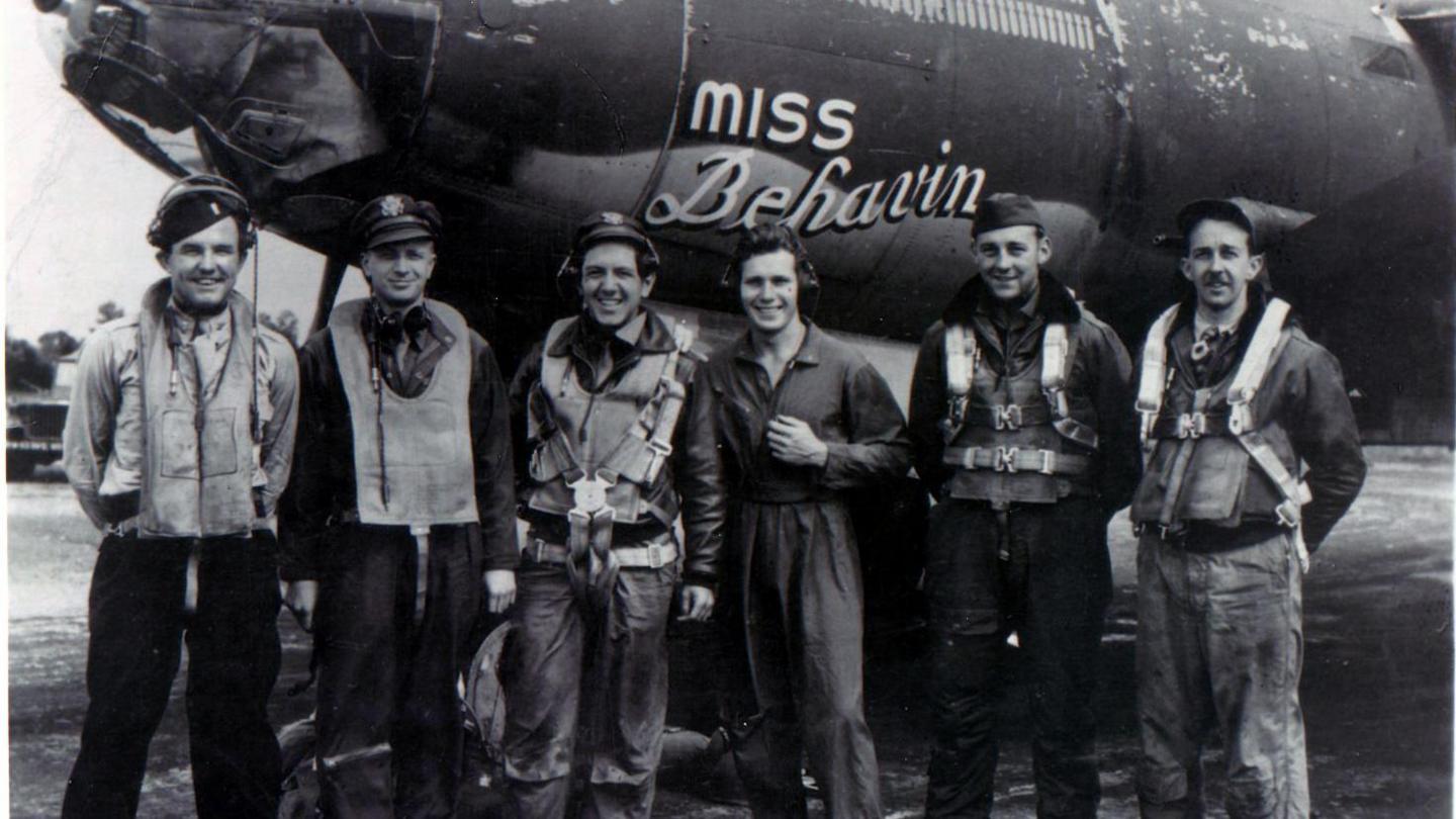 A black and white image of six men standing in front of an aircraft named "Miss Behavin". They are smiling.