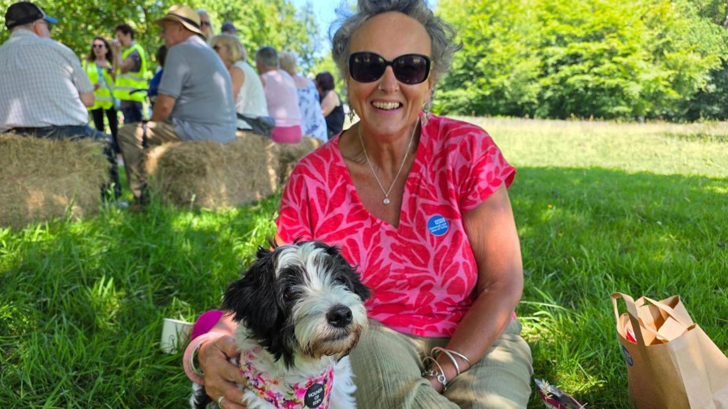 Jennie Brown with her dog sitting on a grass bank. She is wearing a pink top and black sunglasses.