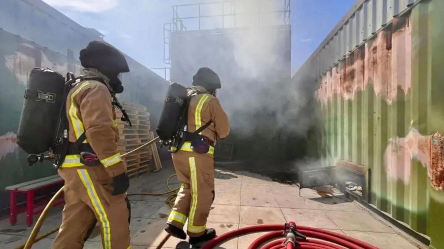 Two firefighters wearing breathing apparatus and yellow protective clothing approach a smoke-filled enclosure surrounded by green and grey metal fencing. A red hose is coiled on the ground next to them.