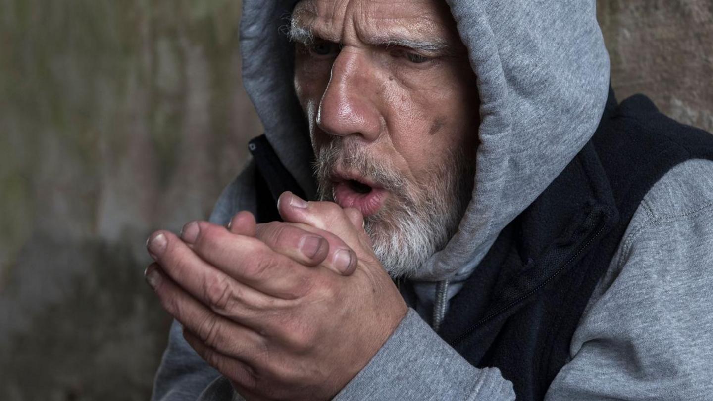 A man with a grey beard rubbing his hands together while blowing air on them trying to keep warm. He is wearing a grey hoody with the hood up and a navy body warmer.