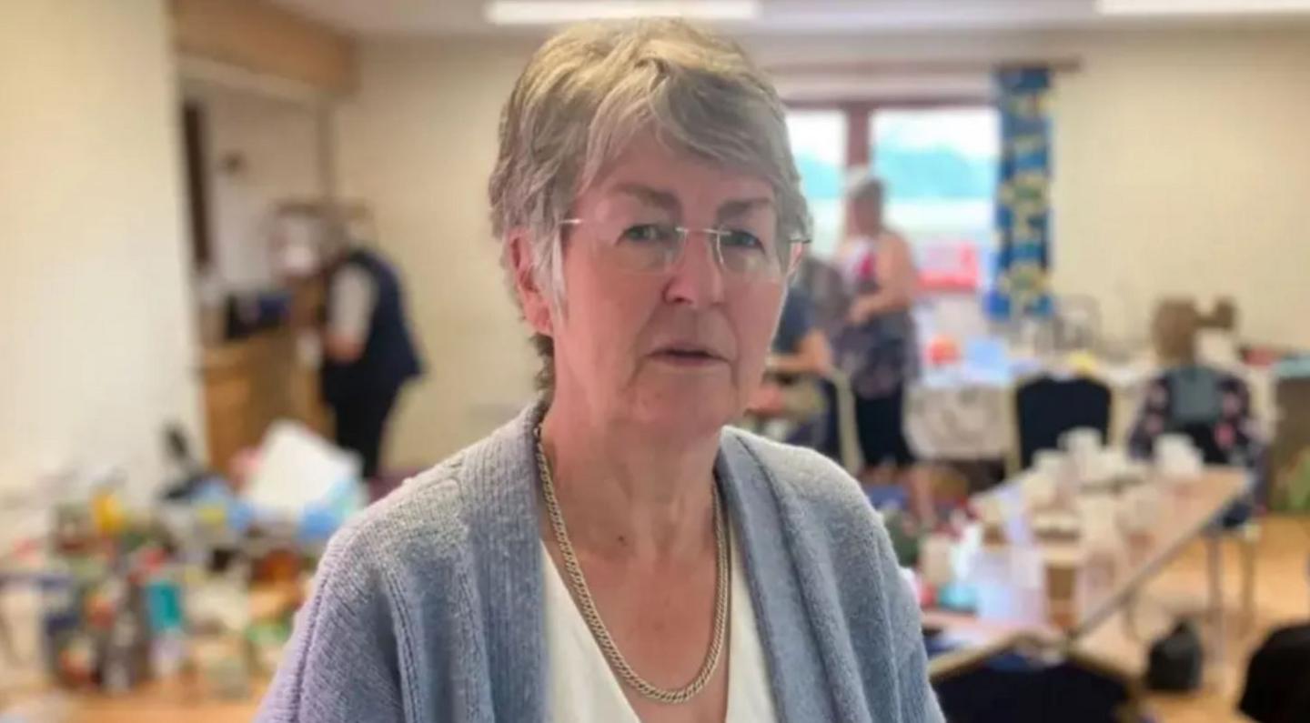 A woman with short grey hair and glasses with a white top and a pale blue cardigan in front of a blurred background